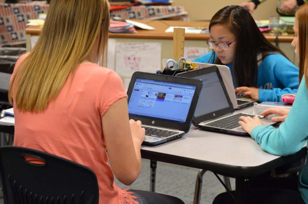 Three students seated at a table using laptops in a classroom setting.