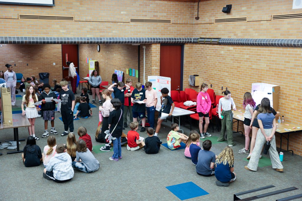 Group of people gathered in a room, some seated on chairs and benches, others standing, all facing towards a large screen
