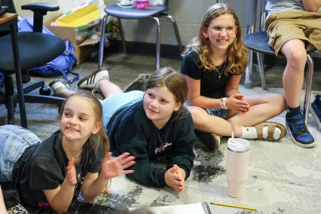 Three young girls sitting on the floor in a classroom setting, smiling and looking at something off-camera.