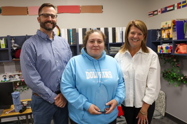 Three people posing in a room with a trophy, a desk with office supplies, and a shelf with books.