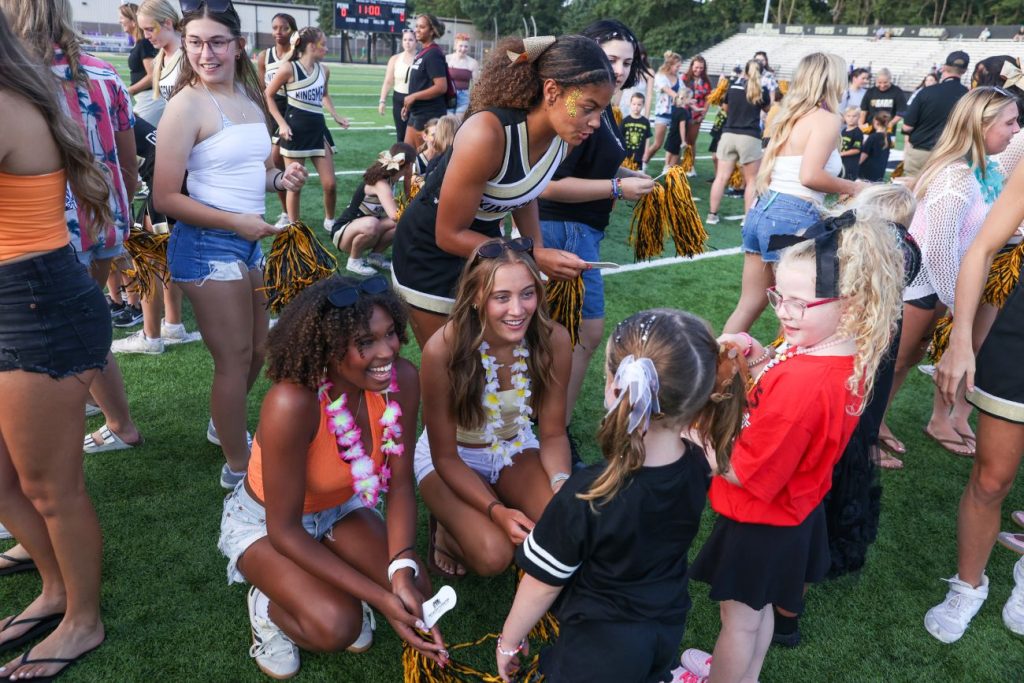 A group of cheerleaders, high school students, and younger students in a stadium setting, with some standing and others sitting on the grass talking and trading pom-poms.